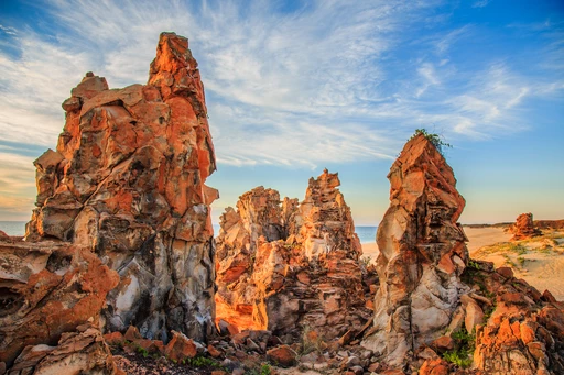 Orangebraune, zerklüftete Felssäulen in dramatischer Landschaft mit Meer und blauem Himmel