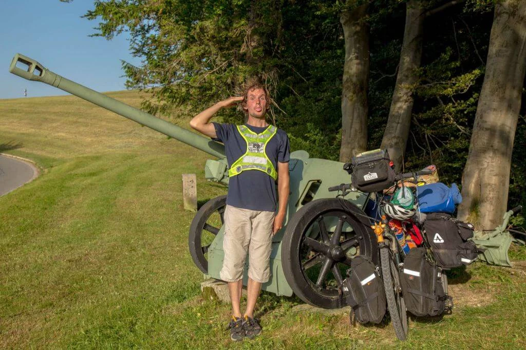 Grüne Militärkanone auf Wiese, daneben eine Person mit Fahrradtaschen und Sicherheitsweste, Bäume im Hintergrund