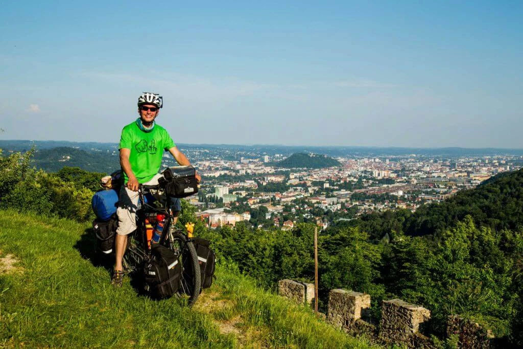 Fahrradreisender in grünem Shirt und Helm auf Wiese, bepacktes Fahrrad, Panoramablick auf große Stadt und bewaldete Landschaft