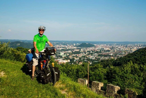 Fahrradreisender in grünem Shirt und Helm auf Wiese, bepacktes Fahrrad, Panoramablick auf große Stadt und bewaldete Landschaft