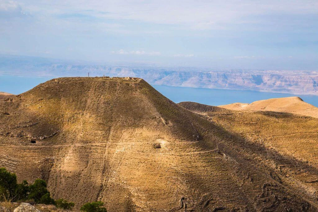 Karges Bergland mit braunen Hügeln und Felshängen, im Hintergrund ein blauer Bergsee und Bergkette