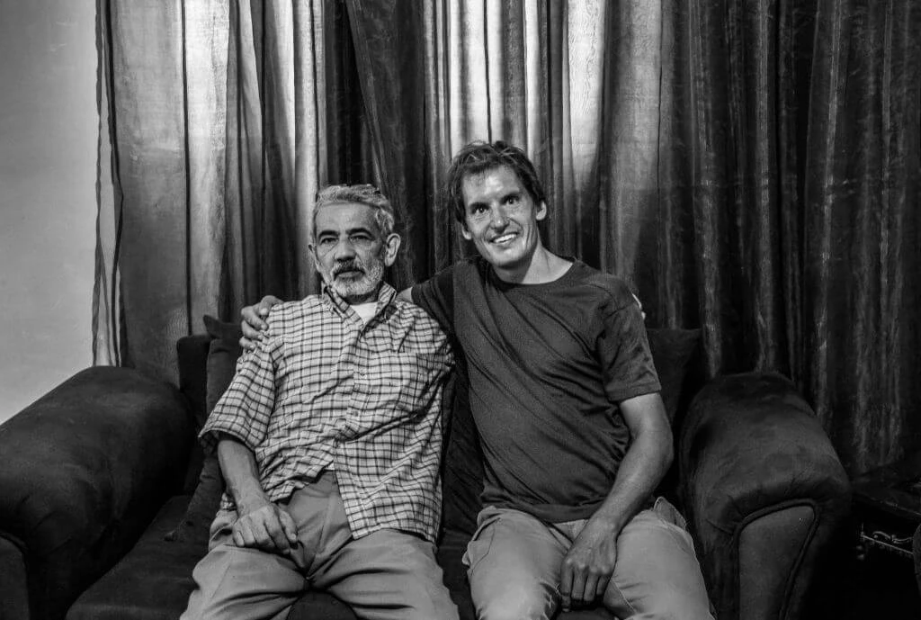 Schwarzweißfoto von zwei Männern unterschiedlichen Alters, gemeinsam auf einem dunklen Sofa sitzend, vor Vorhängen