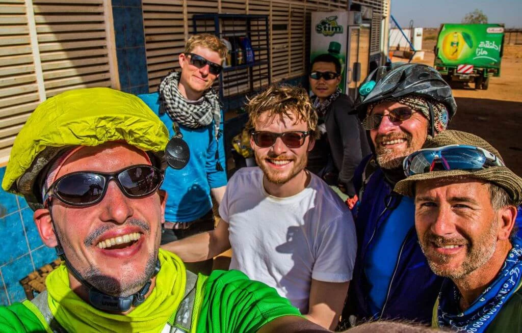 Sechs Männer in Sonnenbrille und Outdoor-Kleidung, Fahrradhelme, lachen zusammen vor Holzwand in trockener Landschaft