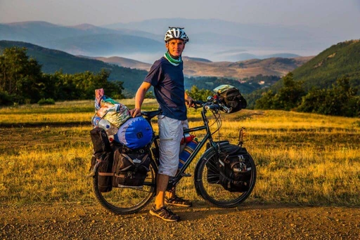 Beladenes Fahrrad mit Packtaschen auf Schotterpiste, Radfahrer mit Helm vor Bergpanorama im Hintergrund