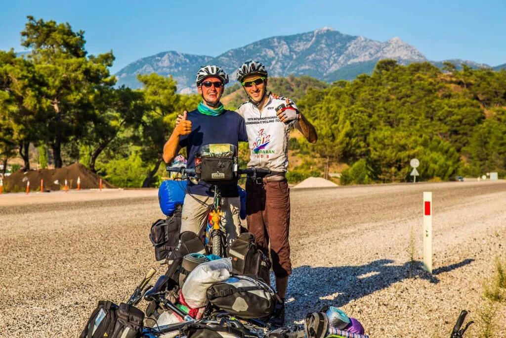 Zwei Radfahrer mit beladenen Fahrrädern auf Schotterweg, im Hintergrund große Berge und grüne Bäume