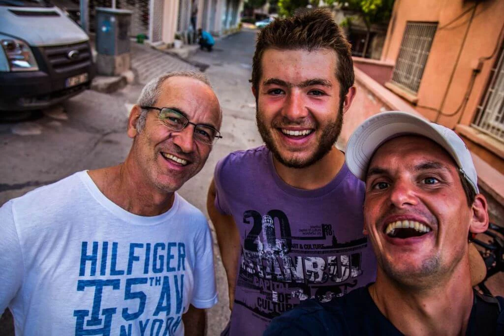 Drei Männer unterschiedlichen Alters stehen auf einer Straße, tragen T-Shirts und lachen herzlich in die Kamera