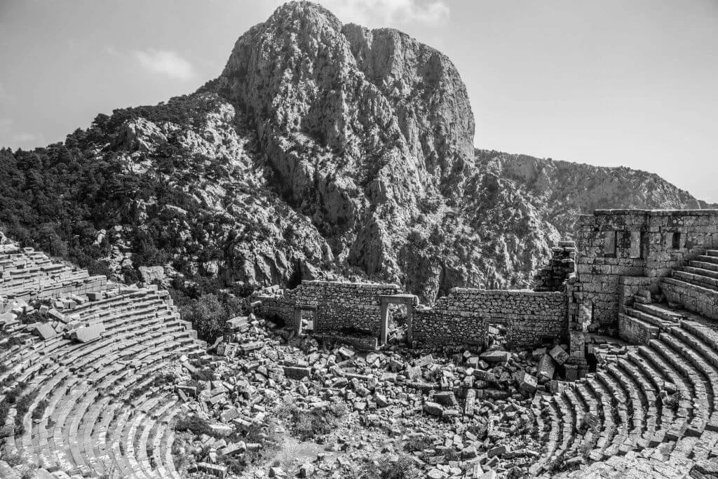 Schwarzweißaufnahme eines antiken griechischen Amphitheaters mit Natursteinstufen, umgeben von zerklüfteten Felsmassiven in dramatischer Berglandschaft