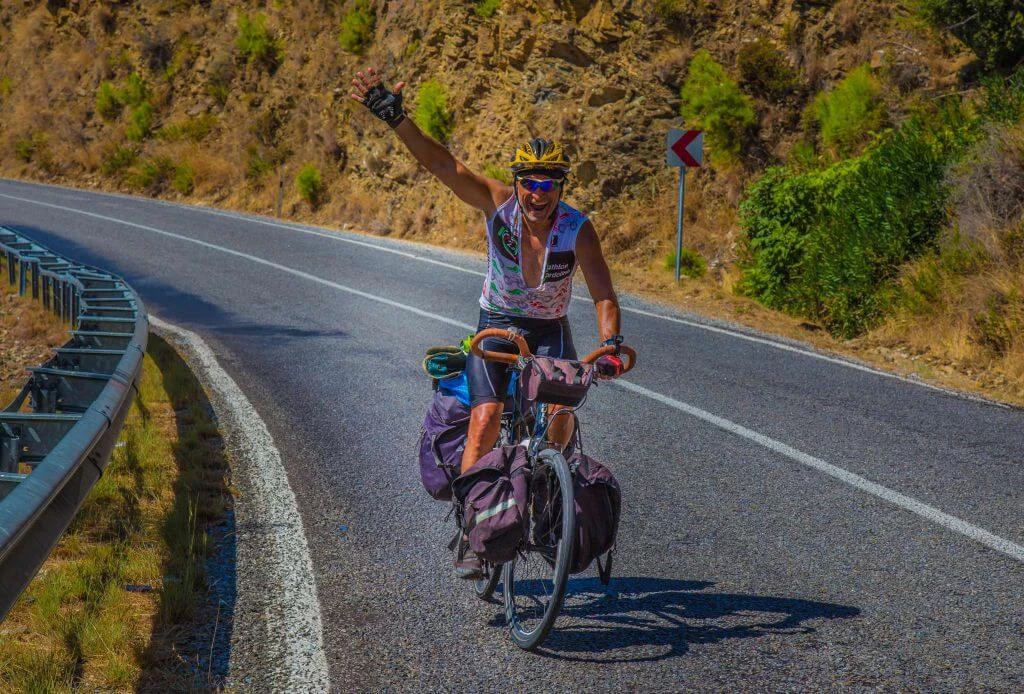 Radfahrer mit Gepäck winkt auf kurviger Bergstraße, trägt Helm und Radtrikot, umgeben von felsiger Landschaft