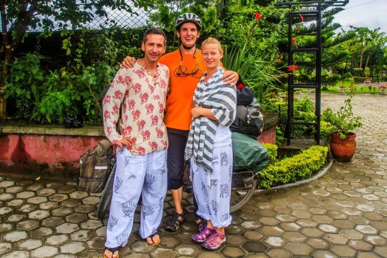 Drei Personen in lockerer Kleidung, einer mit Fahrradhelm, stehen vor üppiger Gartenbepflanzung auf Pflasterweg