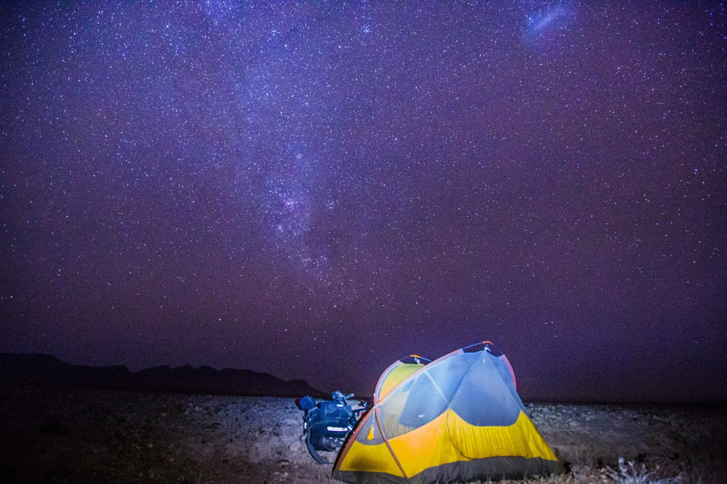 Gelb-blaues Zelt unter einem atemberaubend sternklaren Nachthimmel mit der Milchstraße, felsige Landschaft im Vordergrund