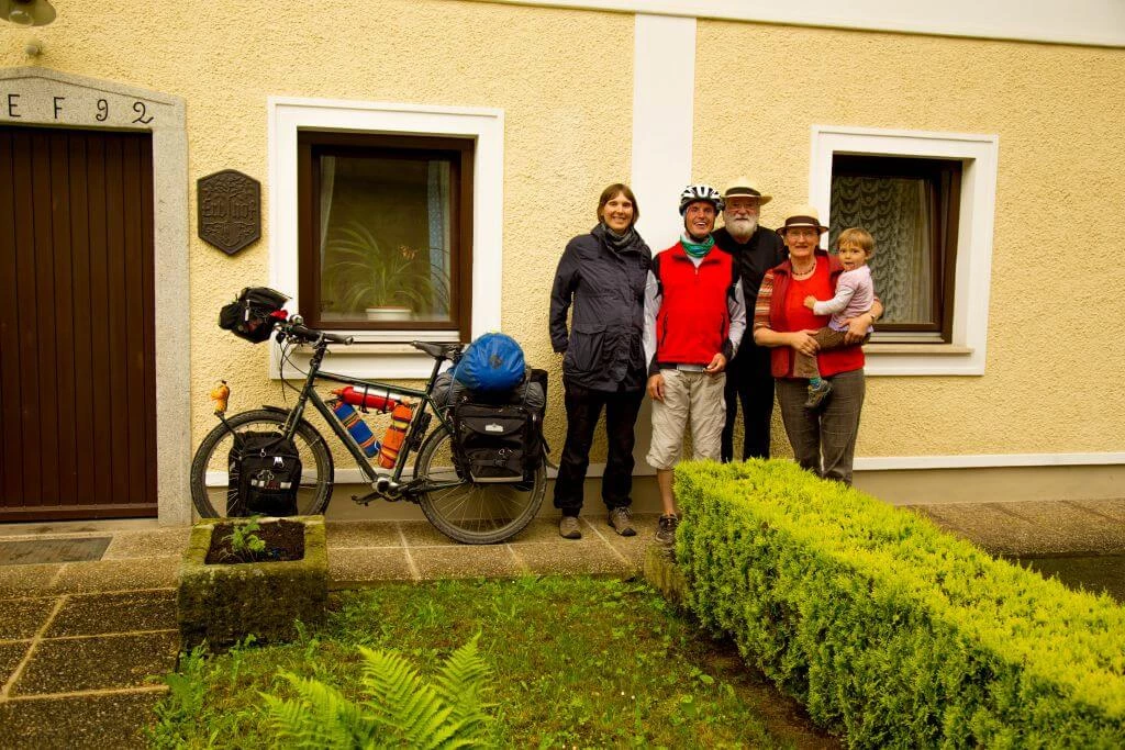 Fahrradreisende Gruppe mit Lastenrad, bestehend aus zwei Erwachsenen, Seniorenpaar und Kleinkind vor gelbem Haus