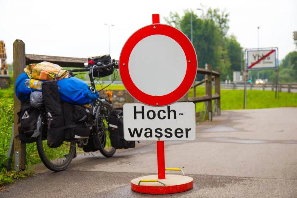 Rotes Verkehrsschild mit 'Hochwasser'-Hinweis blockiert Radweg, beladenes Fahrrad im Hintergrund