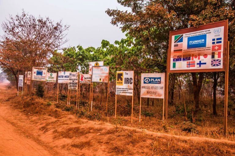 Mehrere Hinweisschilder internationaler Hilfsorganisationen wie UNHCR und IRC an einer staubigen Landstraße mit Bäumen