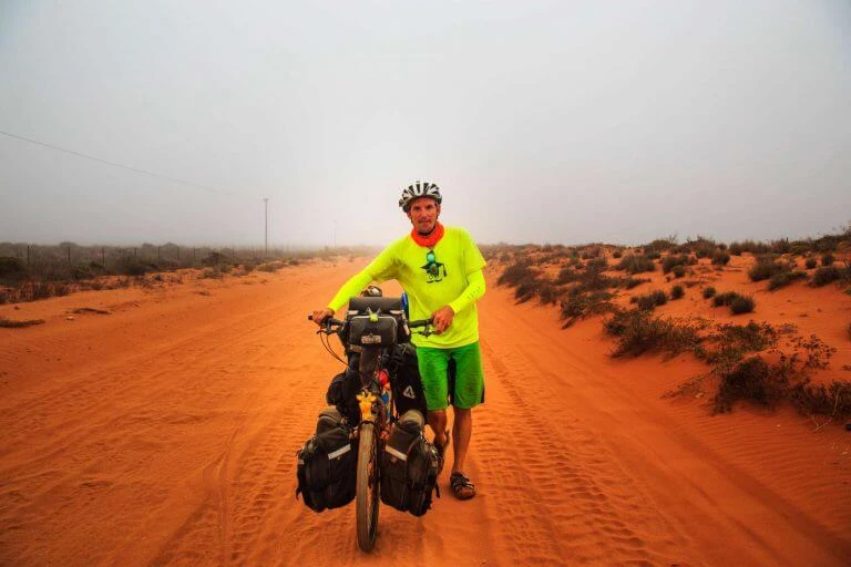 Radfahrer mit gelbem Trikot und Fahrrad auf roter Sandpiste in karger Wüstenlandschaft mit spärlicher Vegetation