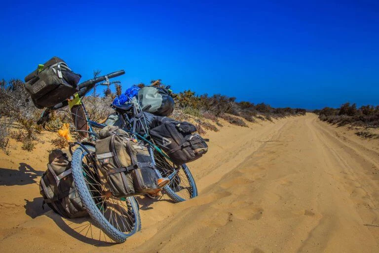 Beladenes Fahrrad mit mehreren Packtaschen auf sandiger Wüstenstraße, blauer Himmel und Sträucher im Hintergrund