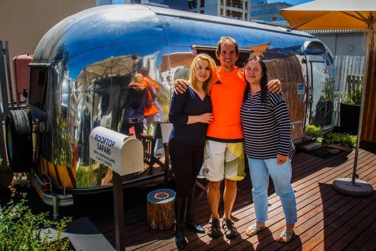 Drei Menschen stehen vor einem glänzend blauen Airstream-Trailer auf einer Holzterrasse an einem sonnigen Tag