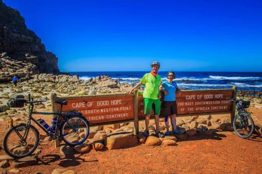 Zwei Radfahrer mit Fahrrädern vor Schild am Kap der Guten Hoffnung, felsige Küste, blaues Meer