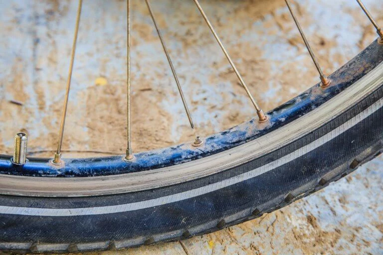 Detailaufnahme eines Fahrradspeichenrads mit blau-schwarzem Reifen, verschmutzt und abgenutzt, auf sandfarbigem Untergrund