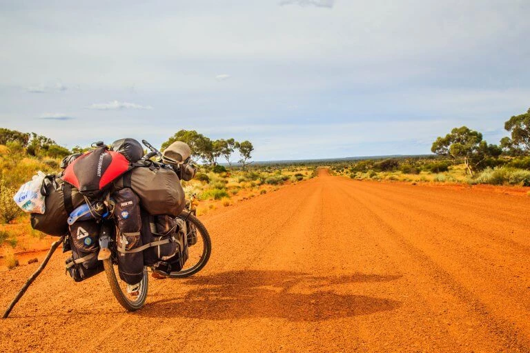 Beladenes Fahrrad auf orangefarbener Schotterstraße, umgeben von Buschlandschaft und blauem Himmel im Outback
