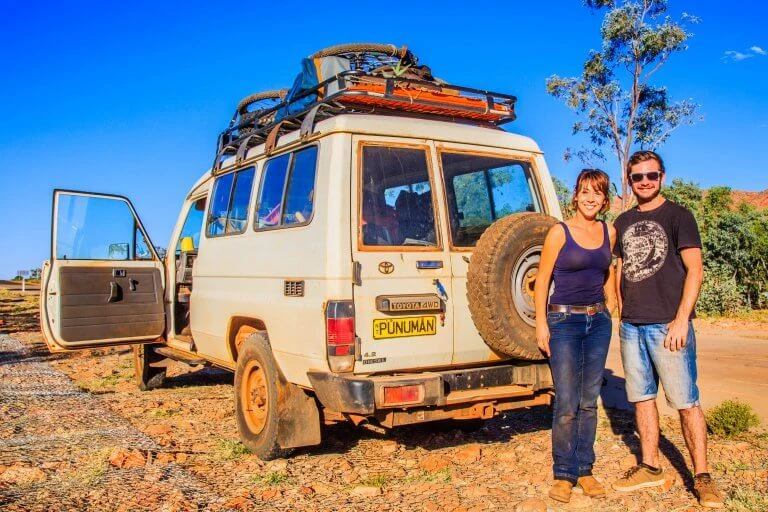 Weißer Toyota Landcruiser mit Dachgepäckträger auf rotem Sandgrund, zwei Personen stehen daneben