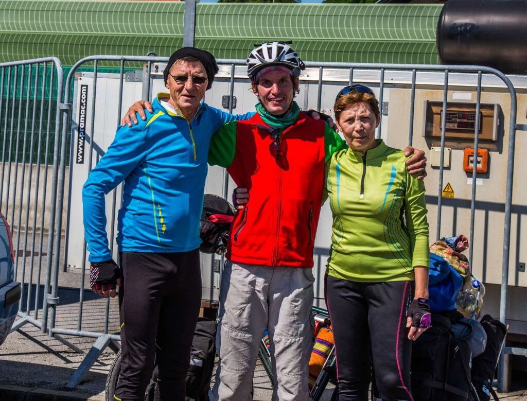 Drei Radfahrer in bunten Jacken - blau, rot, gelb-grün - stehen vor einem Metallzaun und einer grünen Wand