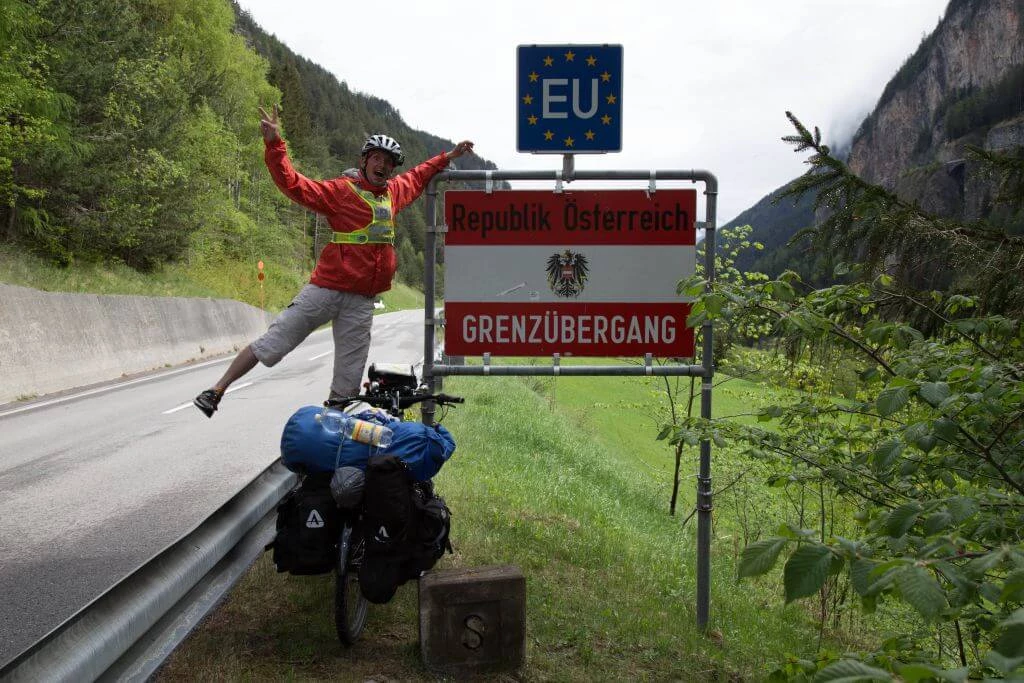 Radfahrer posiert an Grenzschild der Republik Österreich mit EU-Flagge, Mountainbike mit Gepäck, Berglandschaft im Hintergrund