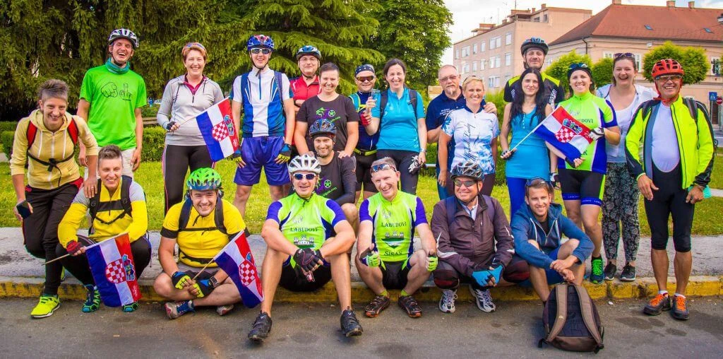 Große Gruppe von Radfahrern in bunten Trikots, mit Fahrradhelmen, versammelt auf einer Straße, mehrere halten kroatische Flaggen
