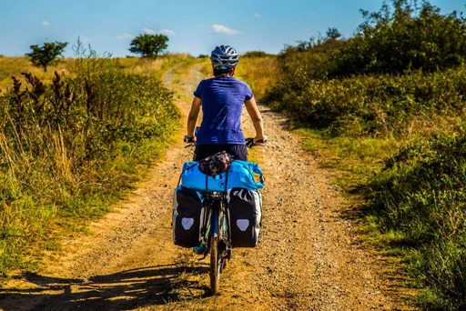 Radfahrer von hinten auf Schotterpiste, blaue Satteltaschen, grüne Landschaft, Sommertag