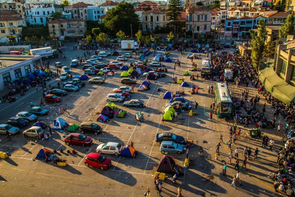 Parkplatz mit parkenden Autos und bunten Zelten, umgeben von Häusern. Viele Menschen in enger Siedlung, Sonnenlicht.