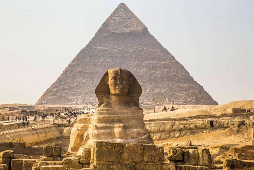 Historische Pyramide und Sphinx aus Sandstein vor blassem Himmel, Besuchergruppen im Vordergrund