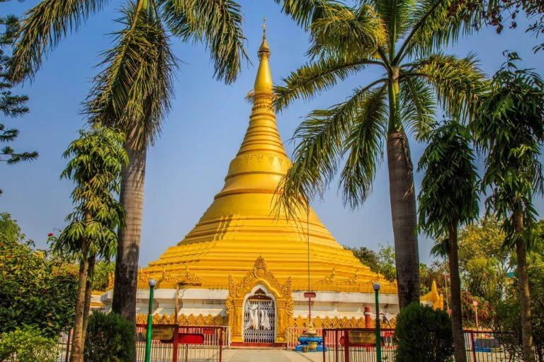 Goldgelbe Pagode mit spitzer Kuppel, umgeben von hohen Palmen vor blauem Himmel, eingezäunter Eingangsbereich