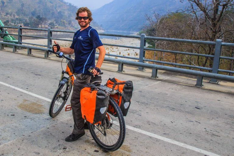 Radfahrer mit beladinem Fahrrad an Bergstraße, blaues Shirt, orange Gepäcktaschen, Bergpanorama im Hintergrund