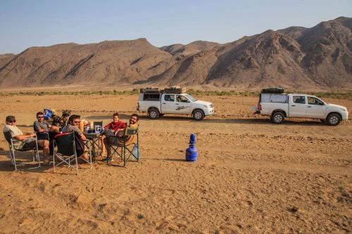 Zwei weiße Geländewagen und Campingstühle in sandiger Wüstenebene mit karger Bergkulisse im Hintergrund