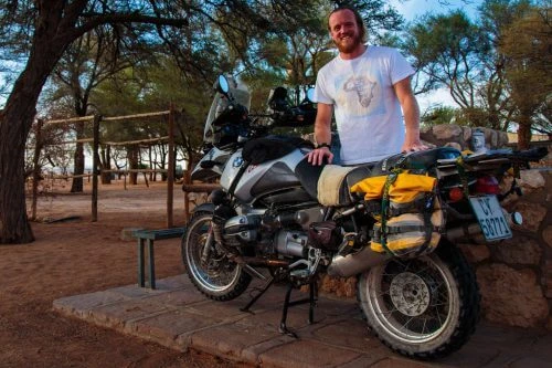 Geparktes Motorrad mit Gepäck unter Bäumen, weißbekleideter Fahrer daneben in sandiger Umgebung