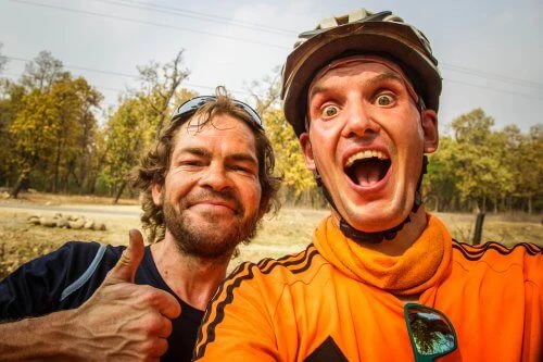 Zwei lächelnde Radfahrer mit Helm in orangefarbener und blauer Jacke im Freien, einer macht Daumen hoch