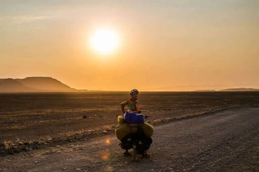 Fahrradfahrer mit Gepäck auf Schotterweg, Sonnenuntergang über karger Berglandschaft im Hintergrund