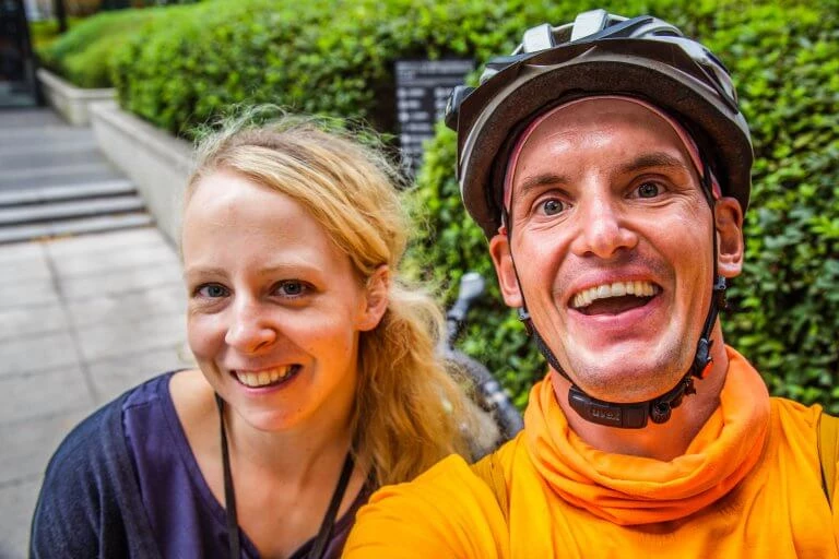 Zwei lächelnde Menschen vor grüner Hecke - eine Frau und ein Mann mit Fahrradhelm in orangefarbener Jacke