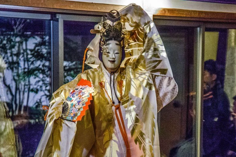Traditionelle japanische Nō-Theatermaske in goldenen Gewändern mit aufwendiger Kopfbedeckung und farbenfächerartigem Accessoire