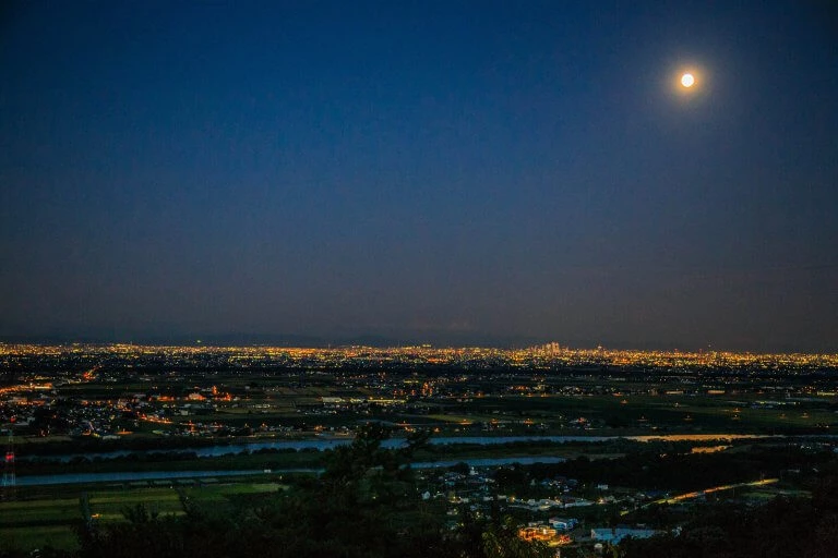 Weite Stadtlandschaft bei Nacht mit beleuchteten Straßen und Gebäuden, darüber heller Vollmond am dunkelblauen Himmel