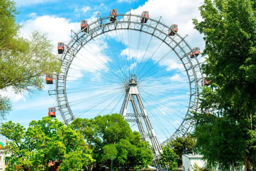 Historisches Riesenrad aus Metall mit roten Gondeln vor blauem Himmel, umgeben von grünen Bäumen