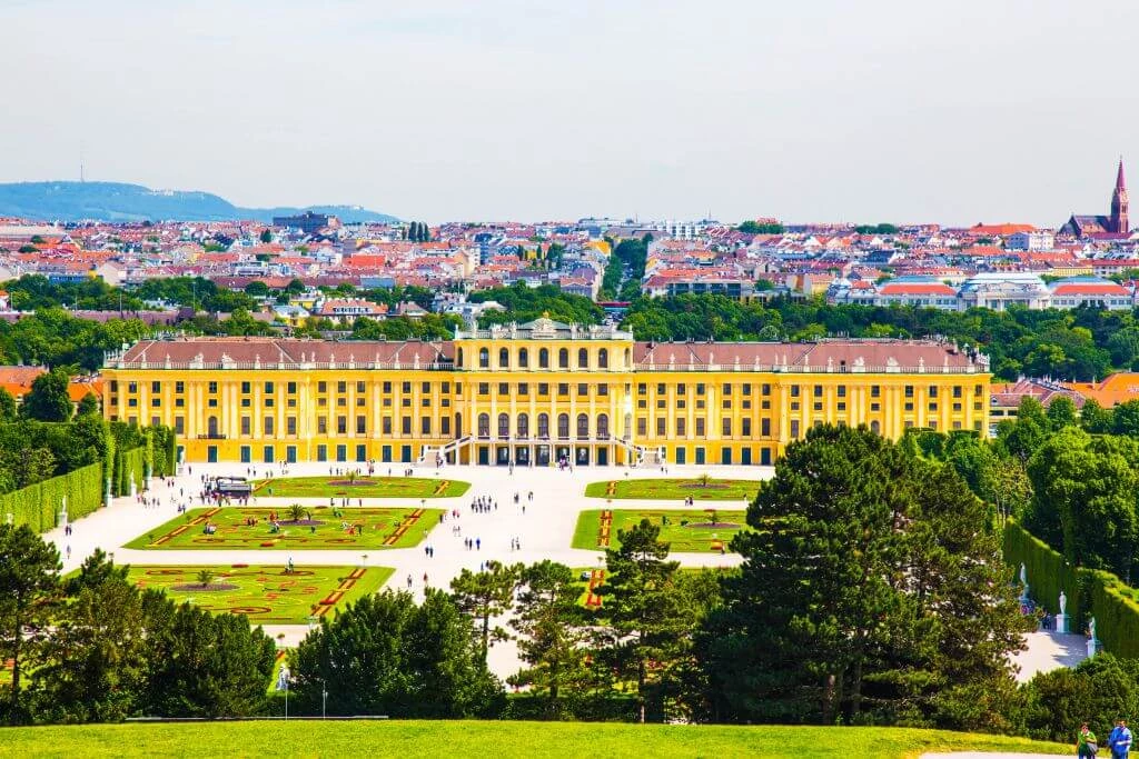 Gelbes Barockschloss Schönbrunn mit symmetrischen Gärten, im Hintergrund Wiener Stadtpanorama und Berge