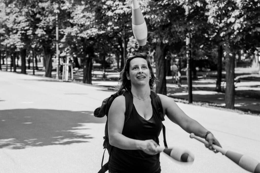 Frau in schwarzem Top jongliert mit weißen Keulen auf einem Waldweg, lächelnd und konzentriert