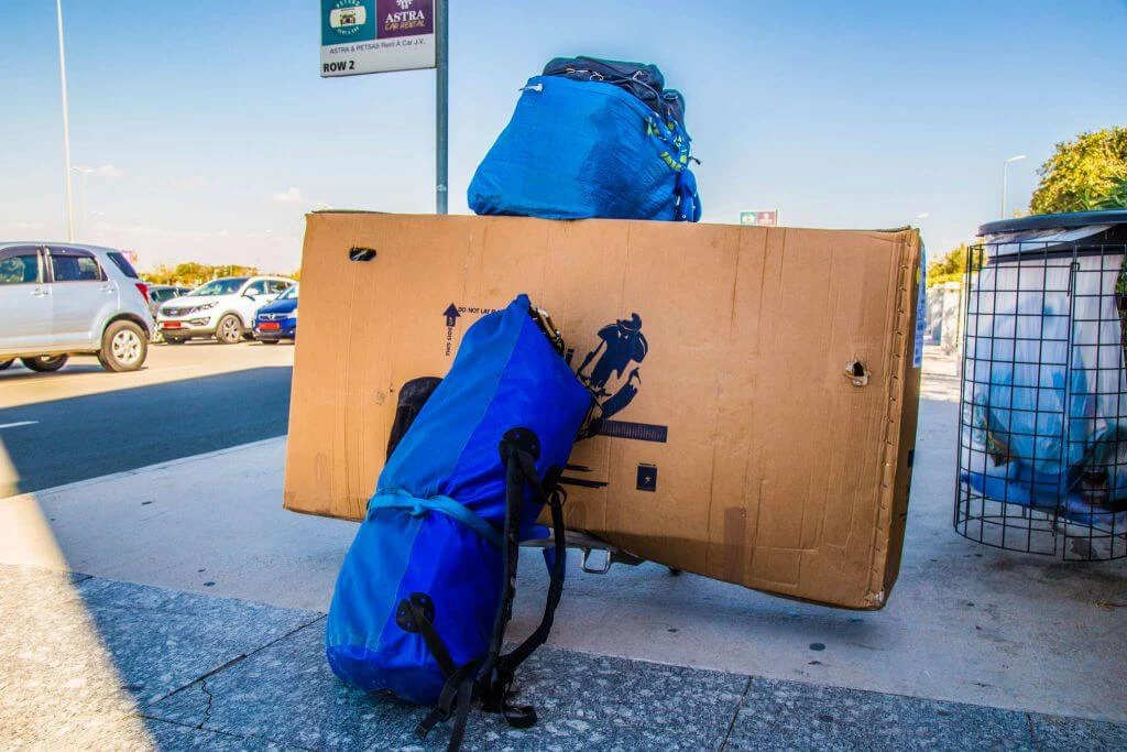 Blaue Fahrradpacktasche lehnt an großem braunem Karton auf Parkplatz mit Astra-Schild und Autos im Hintergrund