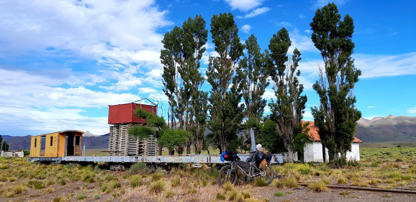 Gelber Eisenbahnwaggon auf Gleis, daneben Fahrrad, umgeben von hohen Pappeln und Berglandschaft mit blauem Himmel
