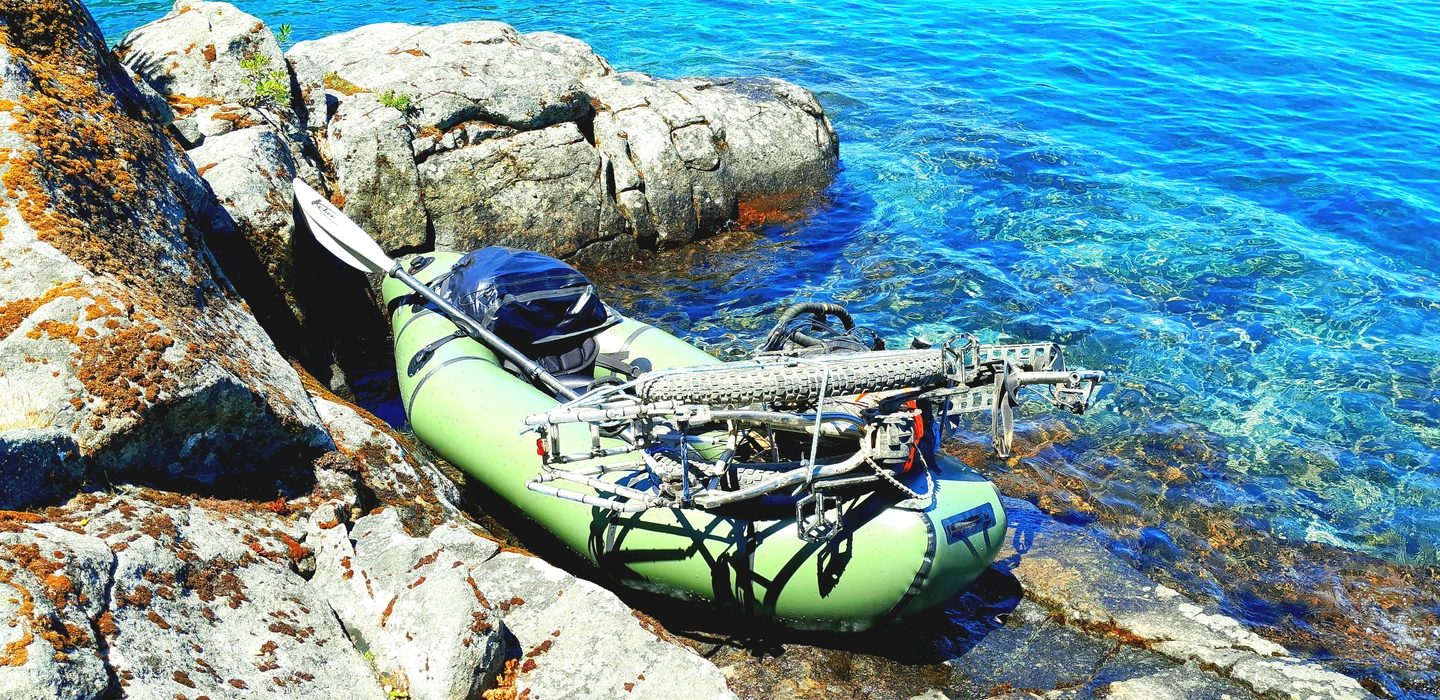 Hellgrünes aufblasbares Packraft mit Metallgestell, vertäut an felsiger Küste mit blauem Wasser und Steinen mit Flechten