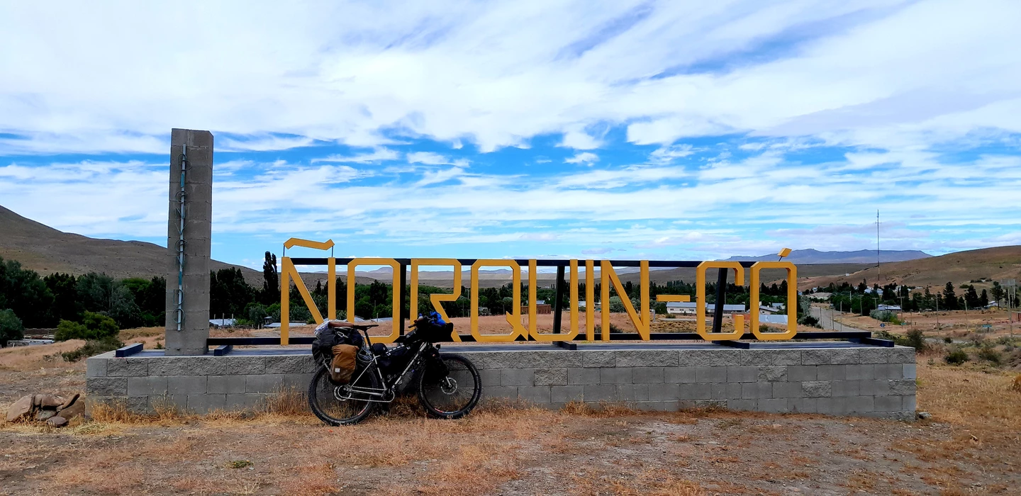 Schwarzes Fahrrad vor gelbem Ortsschild 'Norquinco', Patagonische Landschaft mit Hügeln und blauem Himmel