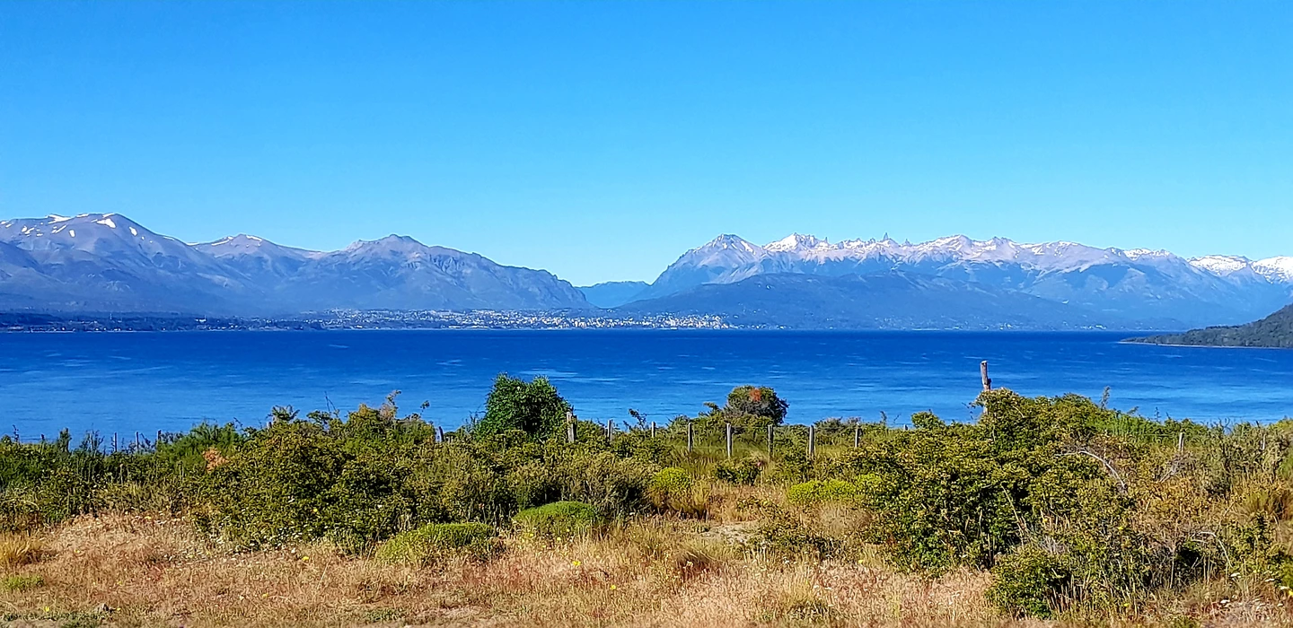 Weite Landschaftsaufnahme mit blauem See, grüner Vegetation im Vordergrund und schneebedeckten Bergen am Horizont unter klarem Himmel