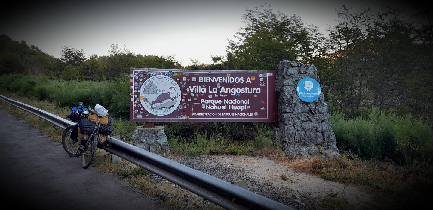 Willkommensschild für Villa La Angostura am Eingang des Nationalparks Nahuel Huapi mit beladenem Fahrrad im Vordergrund