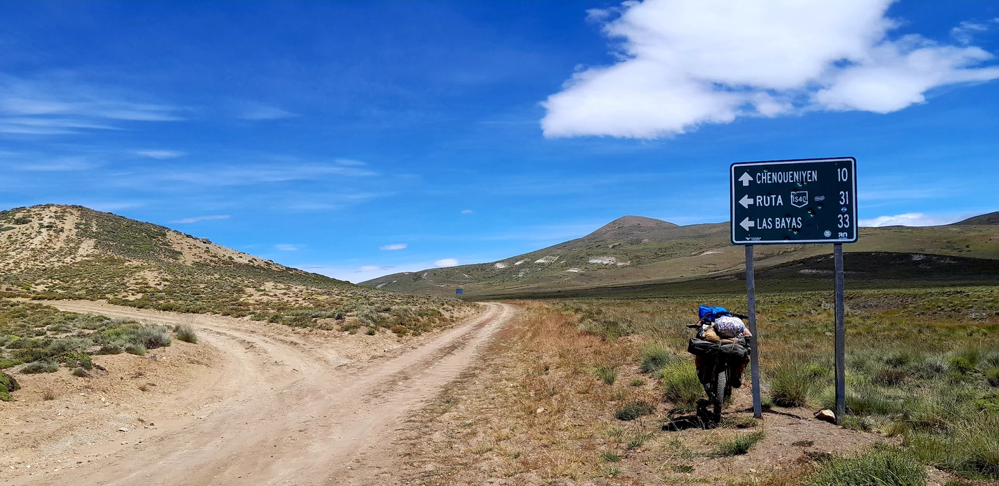 Staubige Schotterstraße gabelt sich in karger Landschaft mit Wegweiser, Bergkulisse und blauem Himmel