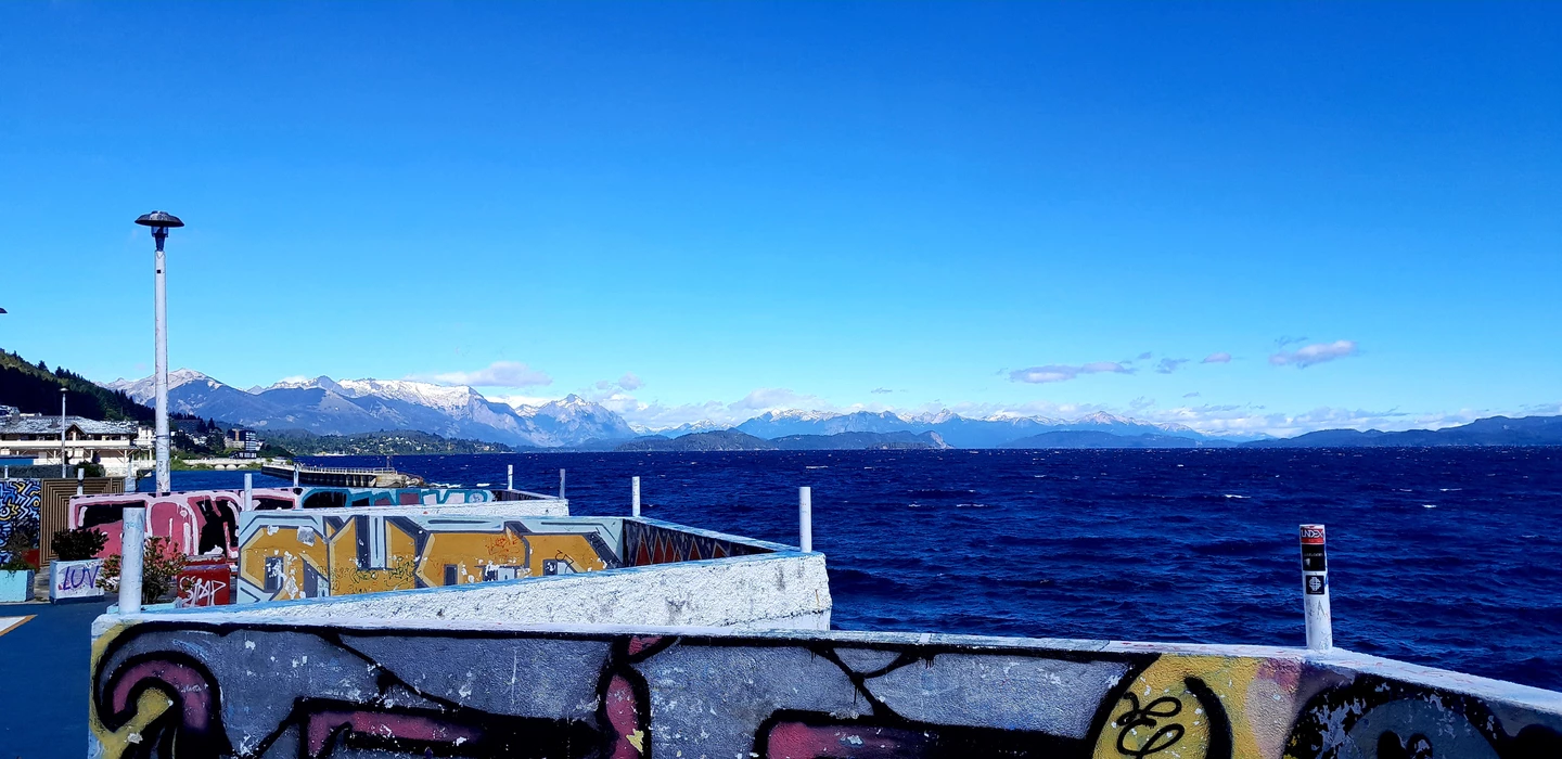 Blick auf eine mit Graffiti bemalte Hafenmole, im Hintergrund blaues Meer und verschneite Berge unter klarem Himmel